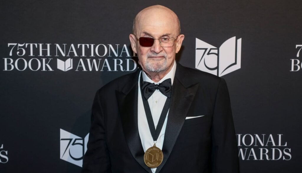 A man encounters a medal around his neck, in front of a backdrop that says "The 75th National Book Award". He has a side of his glasses that was black to cover an eye.